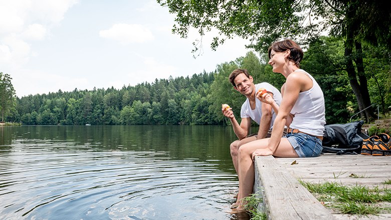 Mann und Frau sitzen auf einem Steg am See und genießen einen Apfel